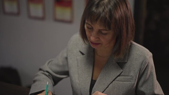 Female caucasian music teacher grading sheet music at desk, marking notation with green pen, grey blazer and notebook visible, certificates on wall, quiet focused school office atmosphere