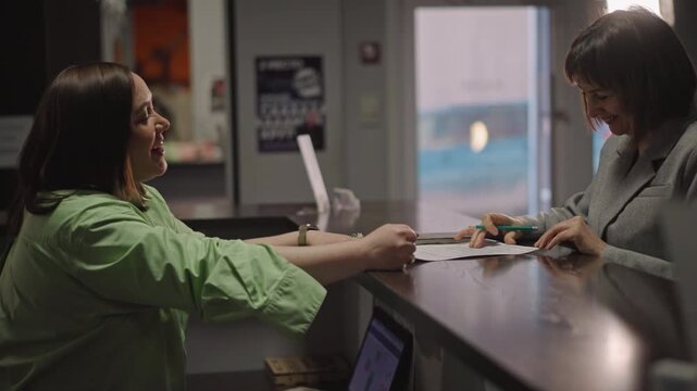 Student handing sheet music at counter during reception interaction, green jacket visible, coach receiving papers and pen, friendly exchange with light through window, procedural yet warm mood