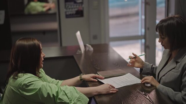 Music school front desk consultation between teacher and student reviewing score and signing lesson registration form, teacher holds pen and points at notation, student leans across polished counter,