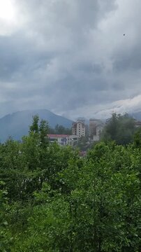 Overcast view of a small town or resort nestled among lush green trees in a mountainous region, with dramatic gray storm clouds moving across the sky and distant snowy peaks