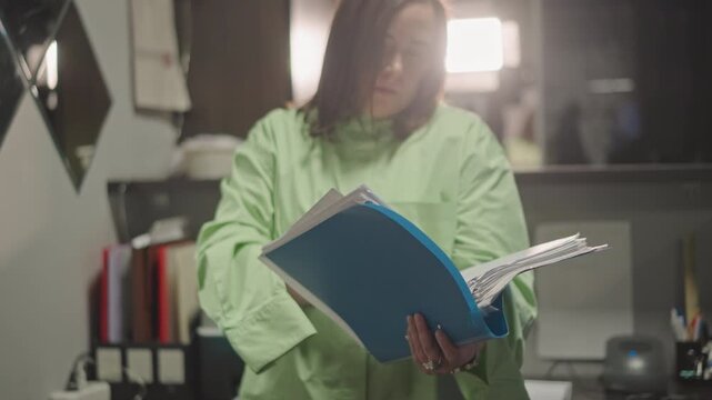 Music coach reviewing blue binder, dramatic backlight and soft lens flare, file shelves and pens visible, turning score pages, concentrated expression, late rehearsal prep, studio desk, focused