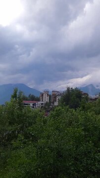 Moody landscape showing a small town nestled on a verdant hillside, with dense forests in the foreground and majestic mountains partially obscured by a dramatic, overcast and cloudy sky