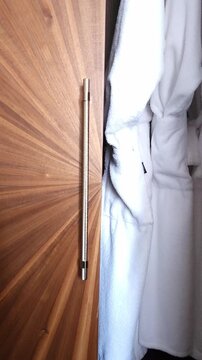 Close up shot of a man's hand opening the doors of a modern wooden wardrobe with a starburst veneer pattern, revealing clean white terry cloth bathrobes hanging inside a hotel or home closet