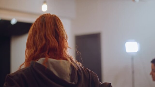Redhead singer facing studio lights, nervous vocalist rehearsing under bright key light, hoodie clad student receives off camera vocal coaching, warm lamp glow, quiet music school atmosphere, close up