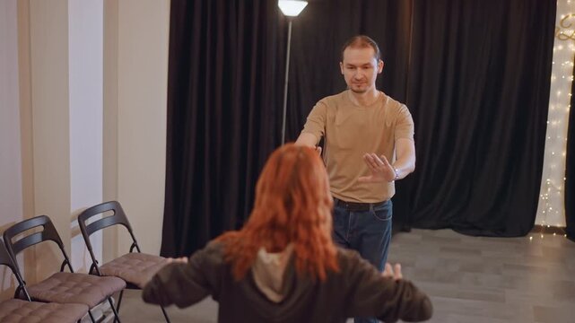 Wide shot white male acting coach guiding student in small blackcurtained studio male instructor uses expressive hands to demonstrate stage blocking and warmup drills rows of folding chairs and soft
