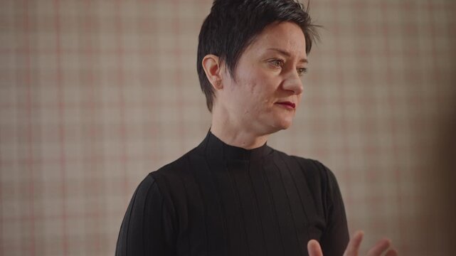 Vocal coach delivering focused lesson in classroom, shorthaired instructor in black sweater critiques student singing, closeup facial expressions, subtle hand gestures, plaid wallpaper backdrop, warm