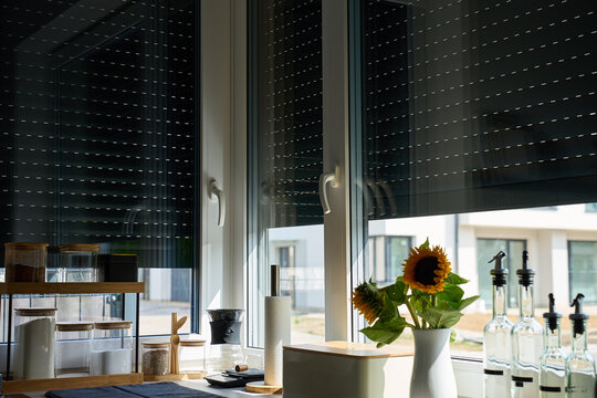 Exterior roller shutters partially cover large kitchen windows above a counter with jars, bottles and sunflowers