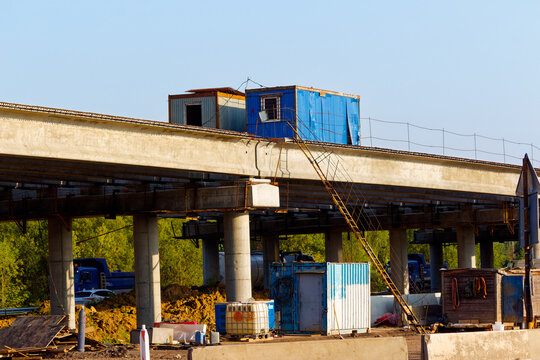Concrete highway overpass under construction featuring industrial containers for workers atop the bridge and temporary site sheds on the ground against a clear blue sky background