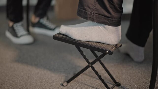 Socked foot on footstool guitar lesson close up of student adjusting foldable footrest while teacher watches in sneakers, soft carpeted floor, warm natural light, focused technique coaching, patient