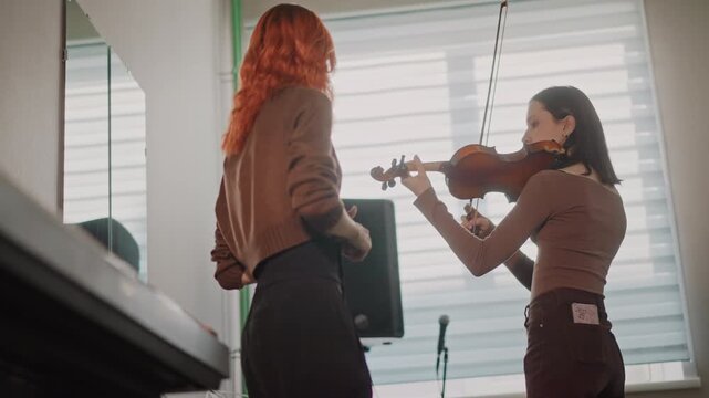 Female musicians practicing duet in bright studio with violin and bow, peer coaching and realtime feedback, speaker and mic setup, focused posture, collaborative energy and recital preparation vibe