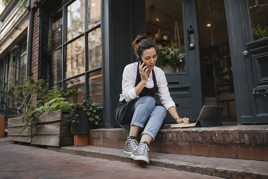 Small business owner managing customer communication on a smartphone while organizing notes beside a tablet outside a local shop, authentic urban entrepreneurship and mobile work.