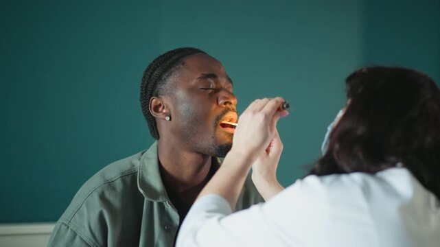 Black male patient receiving oral exam by masked nurse in quiet clinic room, penlight and tongue depressor used to inspect throat and tonsils, calm compassionate interaction, medical assessment