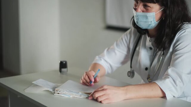 Caucasian doctor taking patient history with pen at desk, masked clinician with stethoscope writing chart entries, attentive listening and calm clinical interaction for accurate assessment