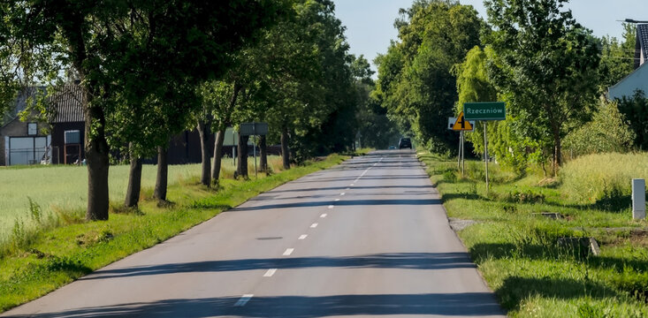  Droga asfaltowa w wojew&oacute;dztwie mazowieckim, a konkretnie w pobliżu Rzeczniowa w Polsce. Droga jest otoczona drzewami i trawą, a nad nią rozciąga się czyste, błękitne niebo. Znak drogowy