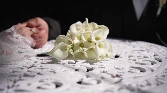 Close-up of a bride and groom holding hands next to a bridal bouquet of white calla lilies, showing an engagement ring.