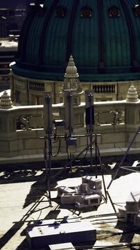 Workers are using equipment at a building site. Structures and tools are visible around the area. The sun is shining, casting shadows of the equipment on the ground.