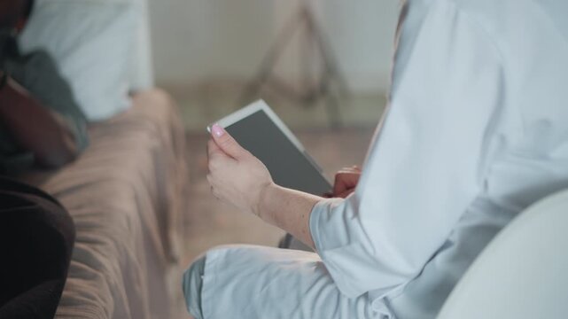 Caucasian caregiver holding tablet beside patient on sofa, gentle interaction in living room. Hands cradle device while scrolling through records, soft ambient light, attentive posture and quiet