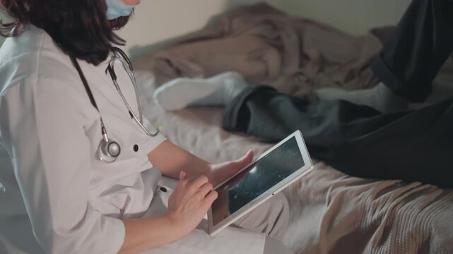White doctor at bedside using tablet, stethoscope draped, reviewing digital records and notes during gentle home visit warm lighting, blanket on patient bed, focused hands tapping touchscreen, calm