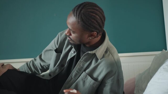 Black man on bed clutching head in pain, tense expression, covers pulled up, sweating and restless, intimate bedroom environment suggesting sudden symptom flare and urgent need for help