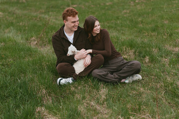 Obraz premium Couple outdoors, relaxed on a grassy field with a friendly goat kid nearby, smiling and affectionate, casual clothing, warm sunlight, gentle breeze, natural colors, peaceful moment, nature lifestyle