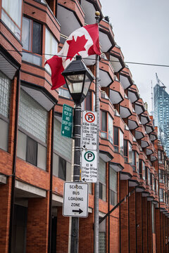 A Canadian flag hangs above american parking, student pick-up and school bus loading signs on a Toronto street, with timed curbside restrictions posted by a red brick residential building in Canada.