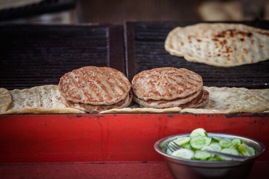 Pljeskavica meat patties cooking on a rostilj stand in Belgrade, Serbia, with grilled flatbread and sliced cucumber visible. The popular grilled meat dish is associated with Serbian and Balkan cuisine