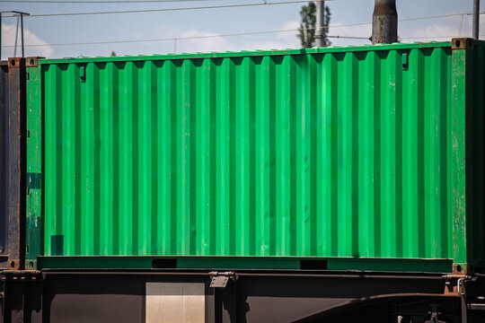 Green corrugated shipping container loaded on a freight train, showing intermodal rail cargo transport, logistics infrastructure and containerized goods movement beneath overhead railway wires.
