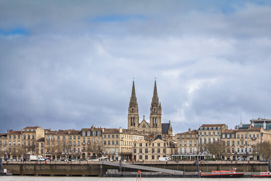 Panoramic view of the Garonne quays in Bordeaux, France, with historic waterfront buildings and Saint Andre Cathedral rising behind the cityscape during a winter afternoon under a partly cloudy sky.