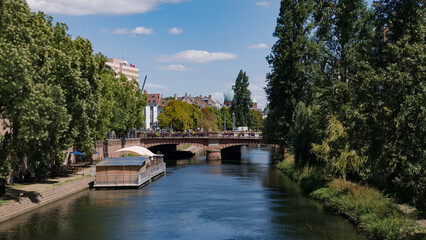Fototapeta premium Ill river flowing past a floating restaurant and bridge in Strasbourg, France, on a sunny day