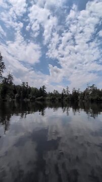 A beautiful, calm lake surrounded by coniferous trees against a backdrop of a blue sky with lovely clouds&mdash;the camera glides across the water's surface; shot from a watercraft. Ontario, Canada
