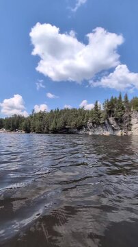 A beautiful, calm lake surrounded by coniferous trees against a backdrop of a blue sky with lovely clouds&mdash;the camera glides across the water's surface; shot from a watercraft.