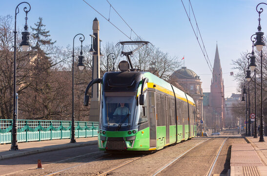 Green modern tram traveling across a historic bridge in central Poznan, Poland, with urban architecture in the background.