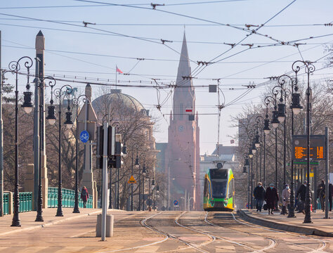 Green modern tram traveling across a historic bridge in central Poznan, Poland, with urban architecture in the background.