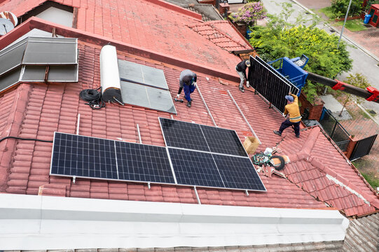Aerial view of workers transporting solar photovoltaic or PV panels with crane onto red rooftop of residential house to install in Malaysia