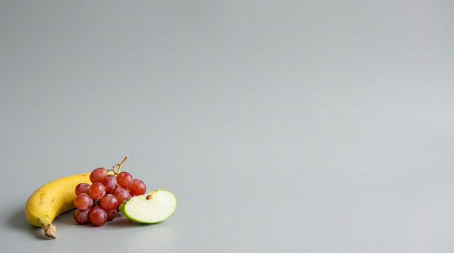 Fresh banana, red grapes, and a slice of green apple on a plain gray background, healthy snack.