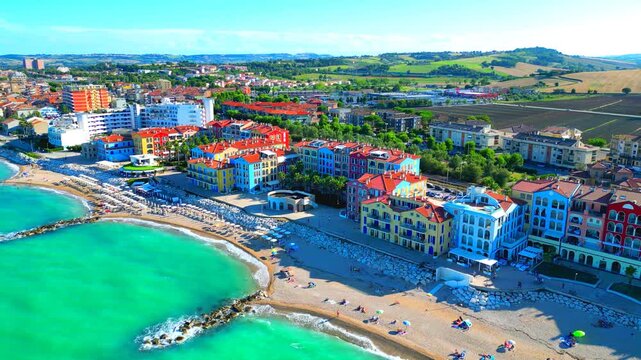 Refreshing aerial truck left pan right arcing shot from Porto Recanati, the aquamarine Adriatic Sea waters gently washing the sandy shore, complete with relaxing bathers, parasols, stones, buildings