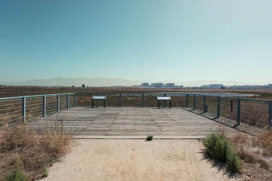 A small wooden observation deck with interpretive signs offers a quiet vantage point over an open salt marsh. Distant office buildings and soft mountain haze fill the horizon beyond the metal railing.