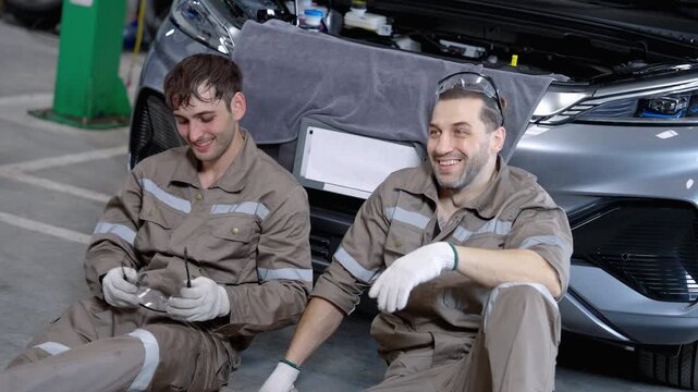 Two automotive technicians wearing protective workwear and gloves take a break beside a car in a professional vehicle repair workshop.
