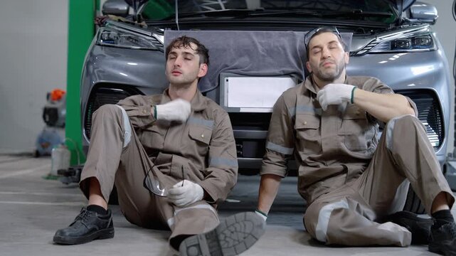 Two automotive technicians in protective workwear sit on the workshop floor and take a break beside a vehicle after completing repair work.
