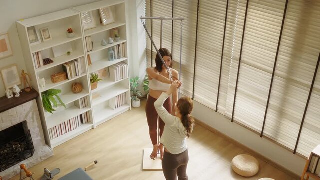 An overhead view captures a pilates instructor guiding her client through a standing suspension ring exercise, both engaged in focused training a dynamic top-down shot of aerial pilates coaching.