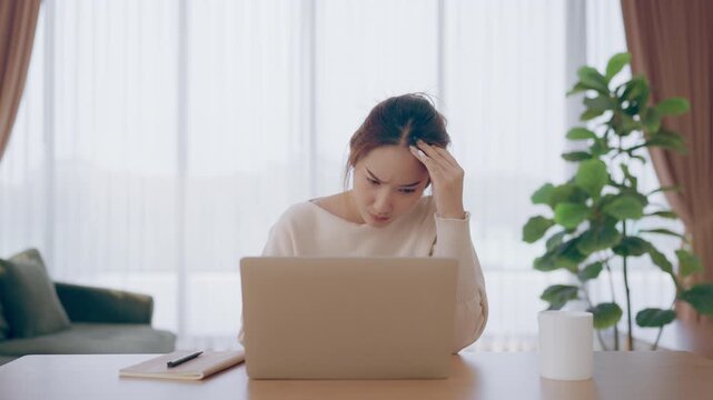 Young Asian woman working in living room, she has a headache due to long and overworking leaving her tired and dizzy. Concept of sickness from work and office syndrome