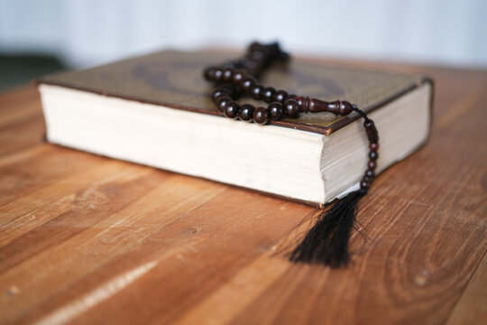 A prayer bead string rests on a closed Quran atop a wooden table; the focus is on the religious object in the foreground against a blurred background&mdash;a concept suitable for Ramadan, Hajj, and Islamic 
