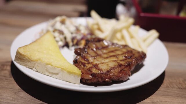 Juicy grilled pork steak with grill marks served on white plate with golden french fries, fresh coleslaw salad, and toasted garlic bread. Delicious restaurant dinner meal on a wooden table.