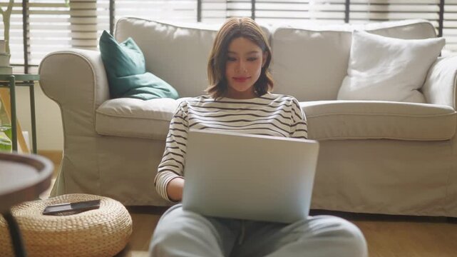 Woman checking smartphone while working on laptop, sitting on floor in cozy living room with plants, vertical remote work multitasking lifestyle scene natural window light	