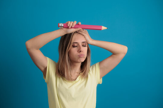 Stressed Young Woman Holding Big Pink Pencil Over Blue Background. Frustrated female student holding a giant pencil on her head making funny face