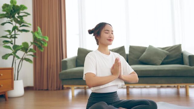 Happy young Asian woman practicing yoga and meditation at home sitting on floor in living room in lotus position and relaxing with closed eyes. Mindful meditation and wellbeing concept