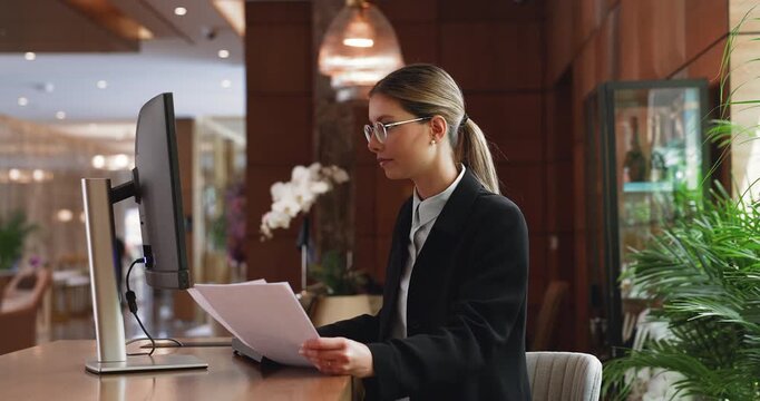 Young woman receptionist in suit sits at reception desk with documents, using hotel software to manage reservations, enters guest information into system during check-in, handling online transactions