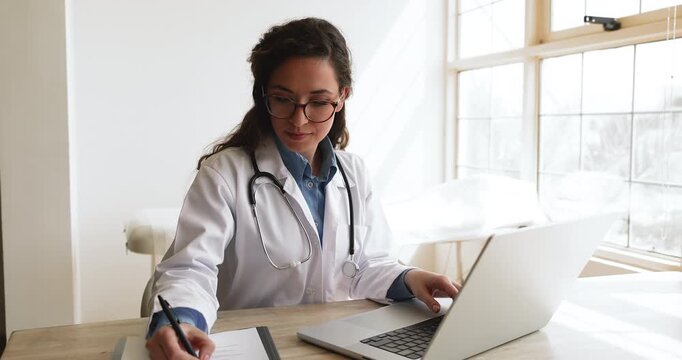 Young Caucasian doctor in glasses and lab coat typing on laptop at workplace, working on medical research, writing notes on clipboard, chatting with patient on telehealth application