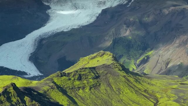 Aerial view of a majestic glacier flowing between vibrant green mossy mountain peaks.