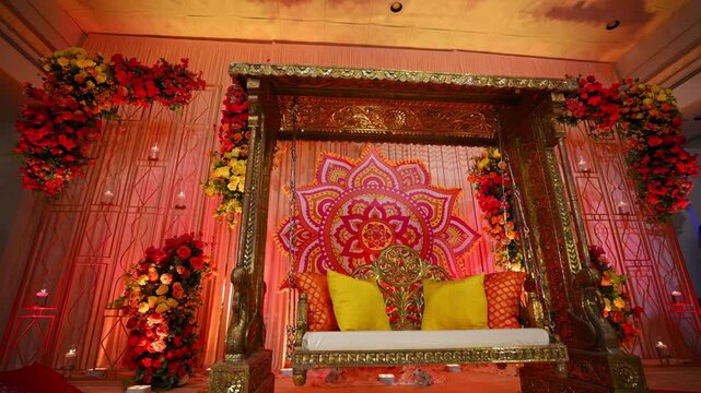 Indian wedding stage decorated with an ornate swing, colorful floral arrangements, mandala backdrop. Traditional ceremonial setup in red, gold, pink, and yellow decorations for a Hindu wedding